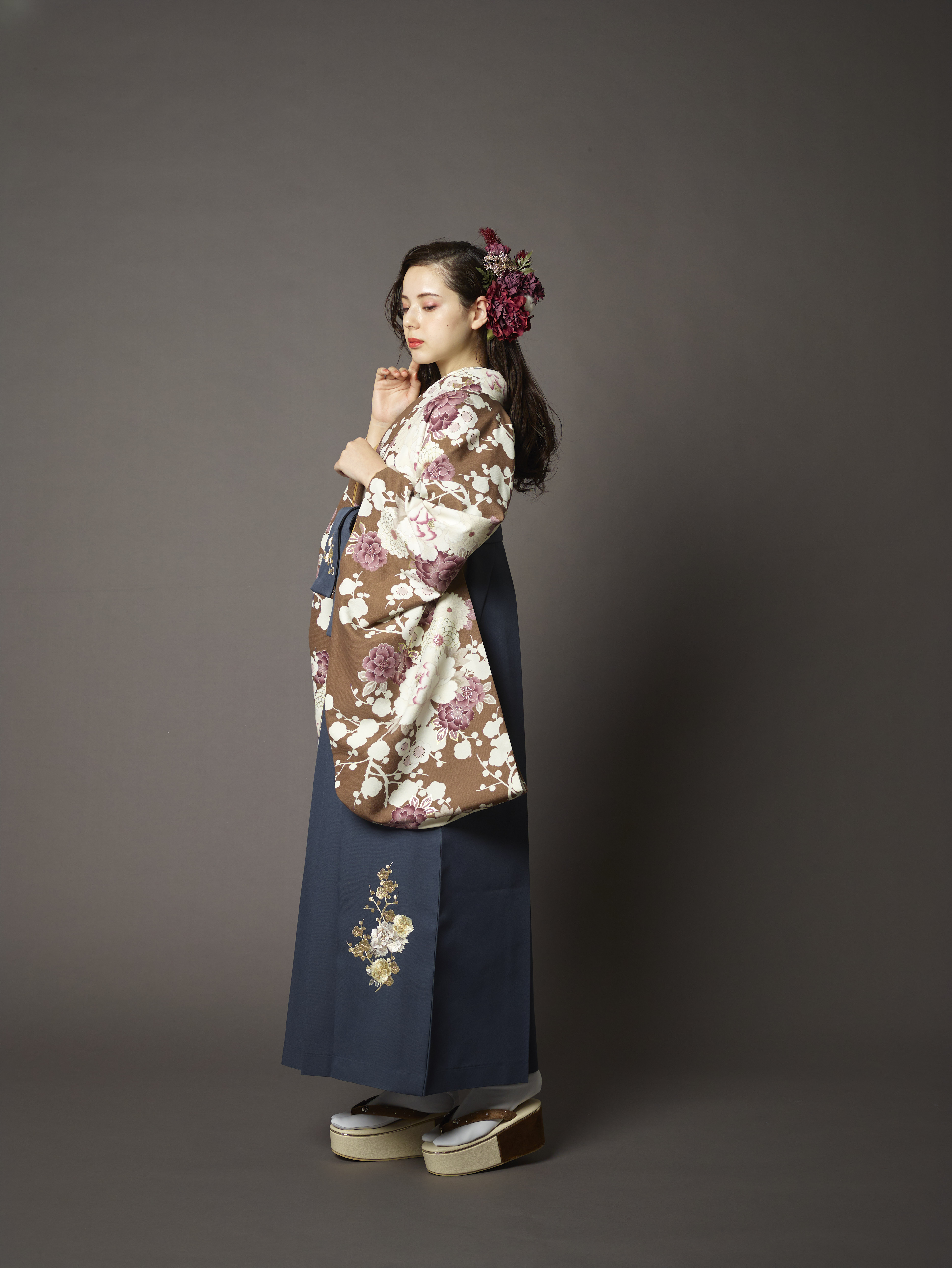 山本寛斎（ヤマモトカンサイ）ブランドの、ブラウン地に白の花柄の二尺袖と、黒の袴の女性用卒業式袴を着用した全身写真